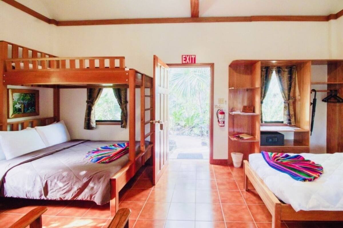 Quarto com beliches, um banco de madeira, uma mesa pequena e uma porta com a inscrição 'EXIT'.