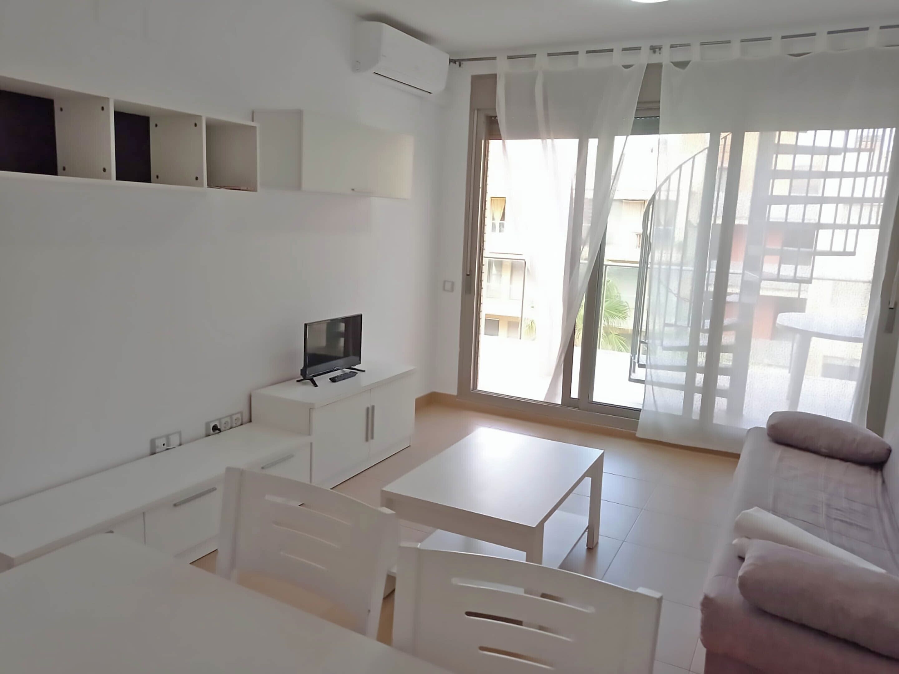 A modern, minimalist living room with a flat-screen TV on a white console, a dining area with a white table and chairs, and a sliding glass door leading to a balcony.