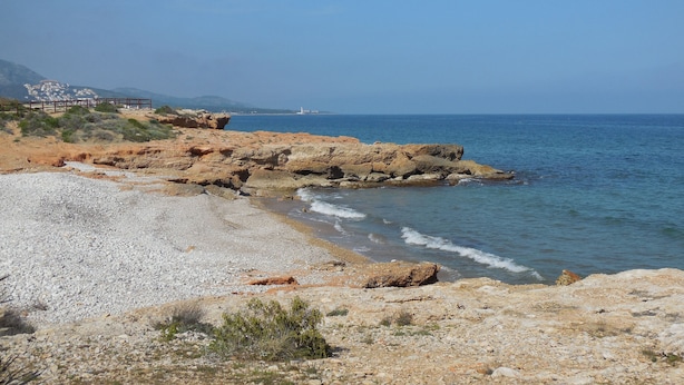 Une plage rocheuse bordée d’une eau d’un bleu limpide, une clôture en bois et des bâtiments qui se dessinent à l’horizon.