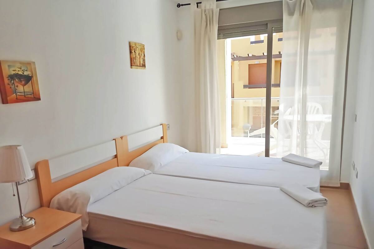 Hotellihuone, jossa on kaksi erillistä sänkyä, yöpöytä lampulla, pieni lipasto ja ikkuna, jossa on verhot.