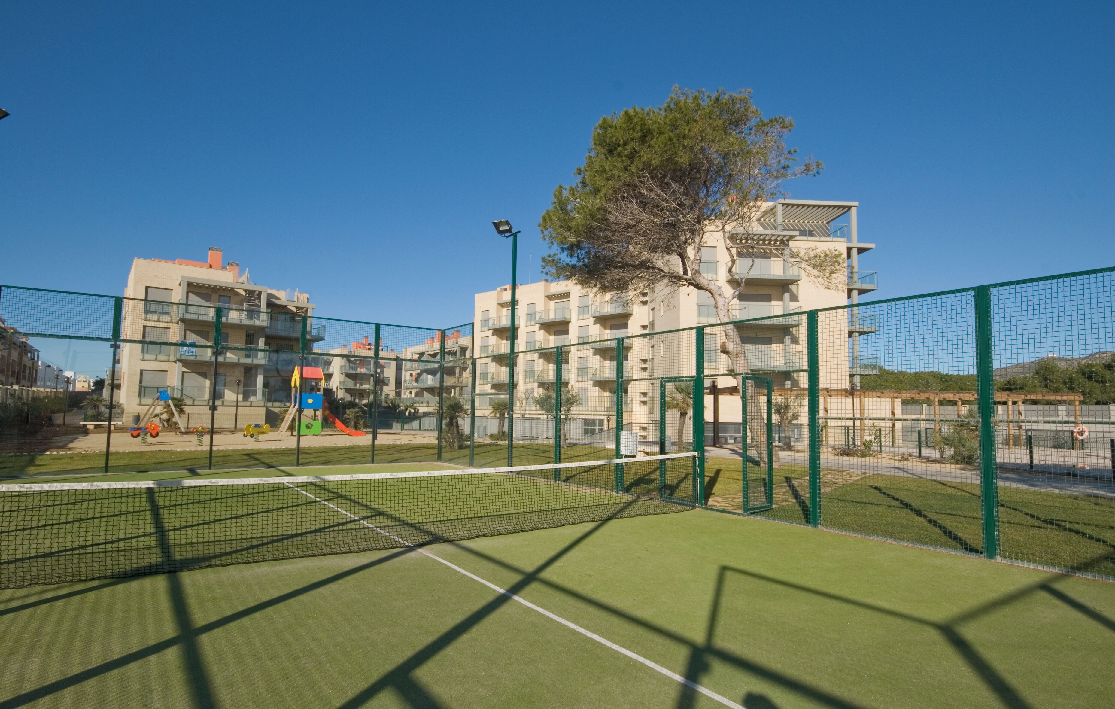 Una cancha de tenis con red, rodeada por una cerca verde y edificios al fondo.