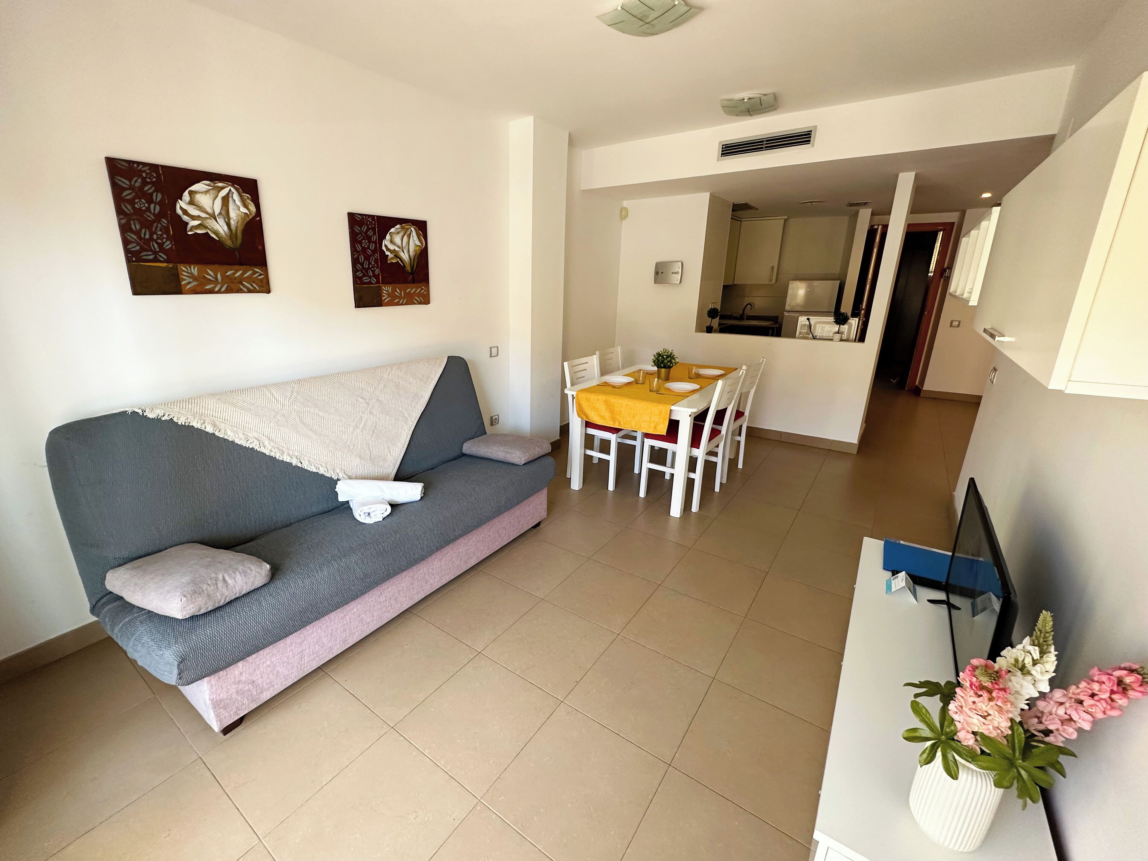 A living room with a grey sofa, a yellow table, white chairs, and a small white vase with flowers.