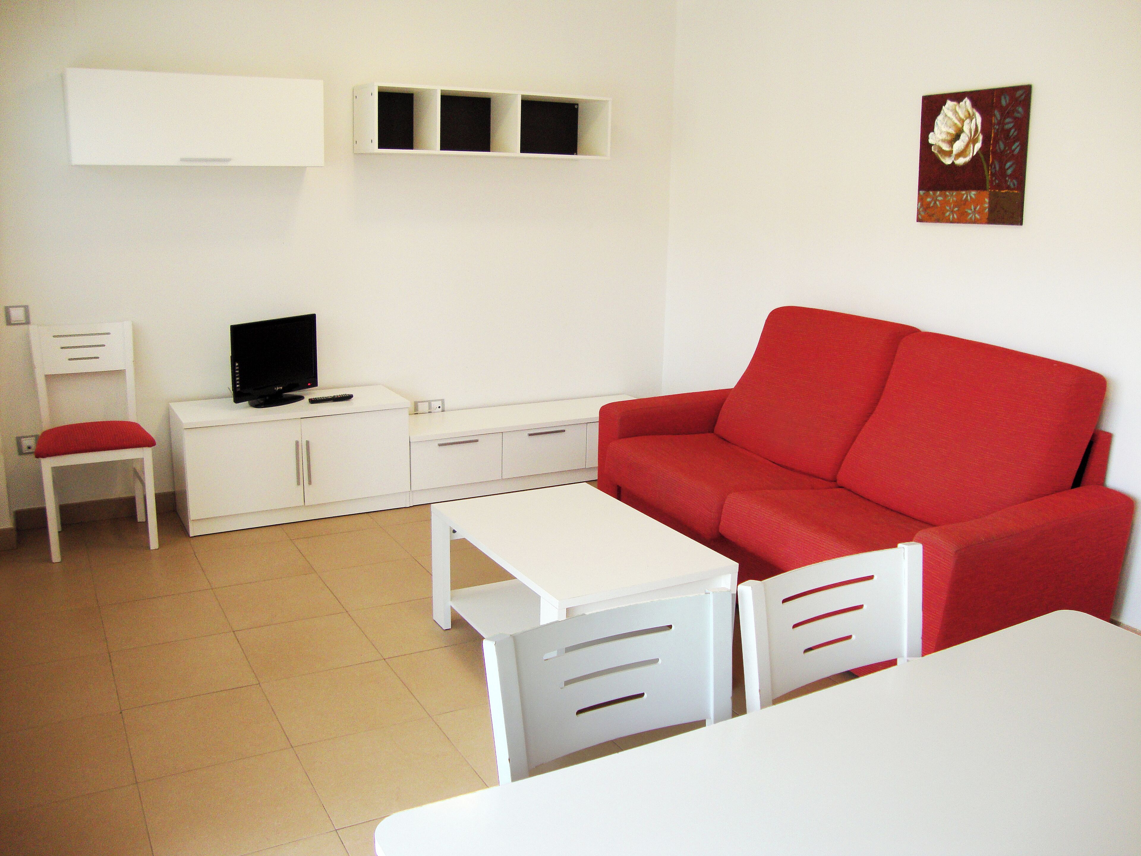 Un petit salon moderne avec un canapé rouge, des meubles blancs, un téléviseur et une étagère fixée au mur.