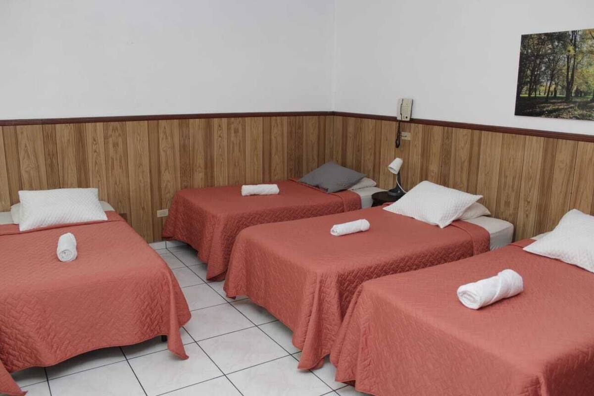Ein Hotelzimmer mit drei Betten, jedes mit einer roten Tagesdecke und weißen Kissen, sowie einem Waldgemälde an der Wand.