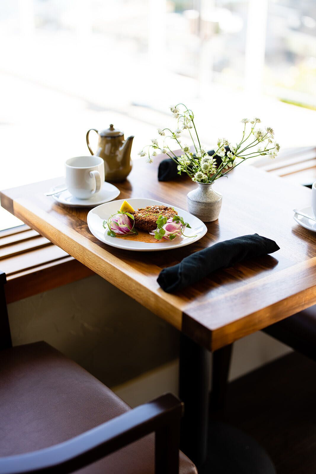 A wooden table with a white plate of food, a teapot, a cup, and a vase with flowers.