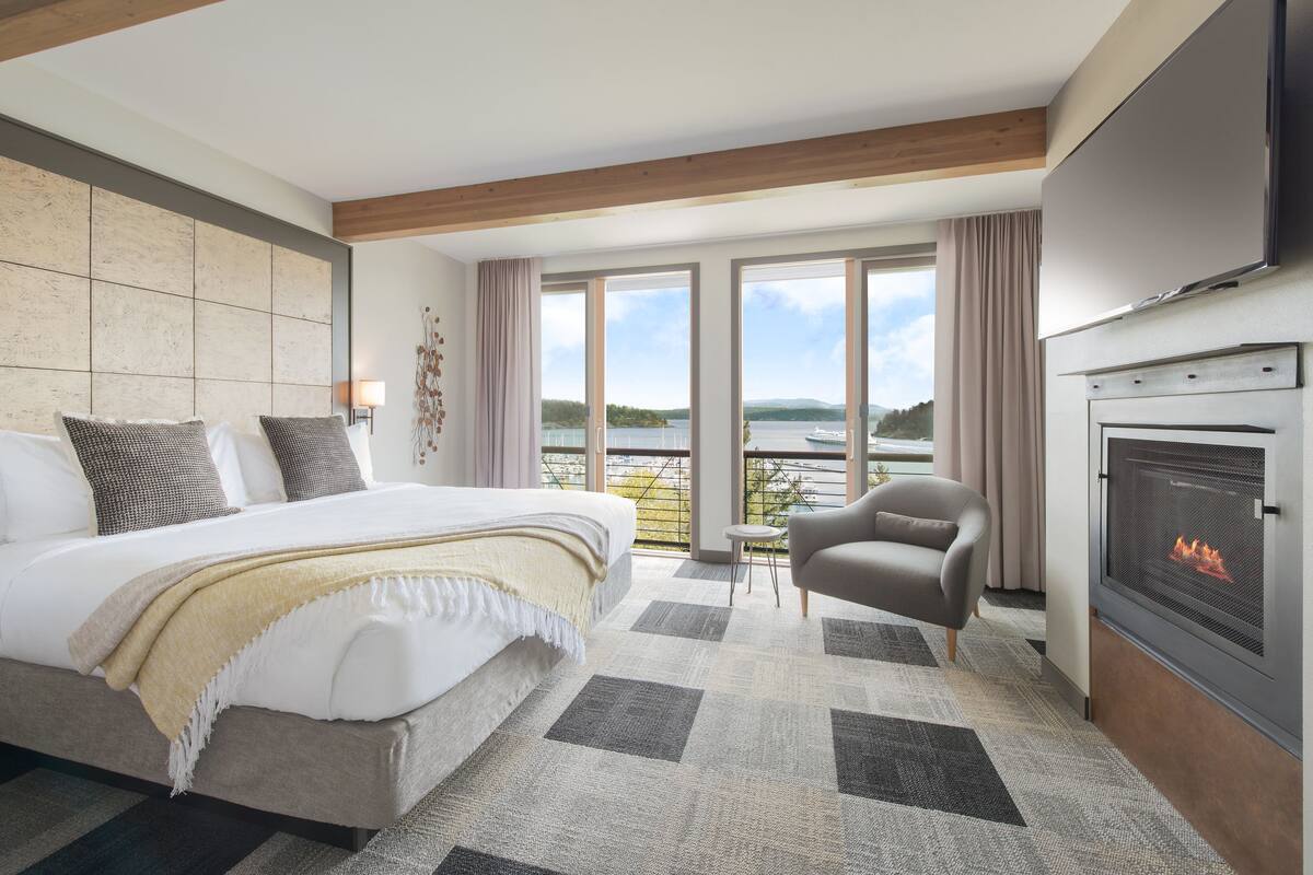 A hotel room with a large bed, a fireplace, a sofa, and a balcony with a view of the lake.