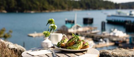 A table with a plate of food, a cup, and a small vase with flowers, overlooking a body of water with boats and docks.