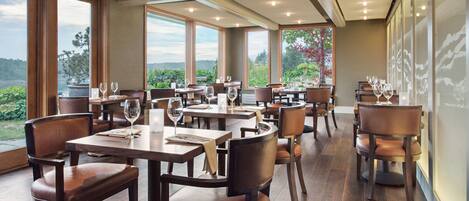 A restaurant with multiple tables and chairs, large windows offering a view of greenery and hills, and a modern interior design.