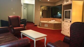 A hotel room with a red carpet, leather chairs, a white table, a TV on a cabinet, and a bathroom with a bathtub and sink.