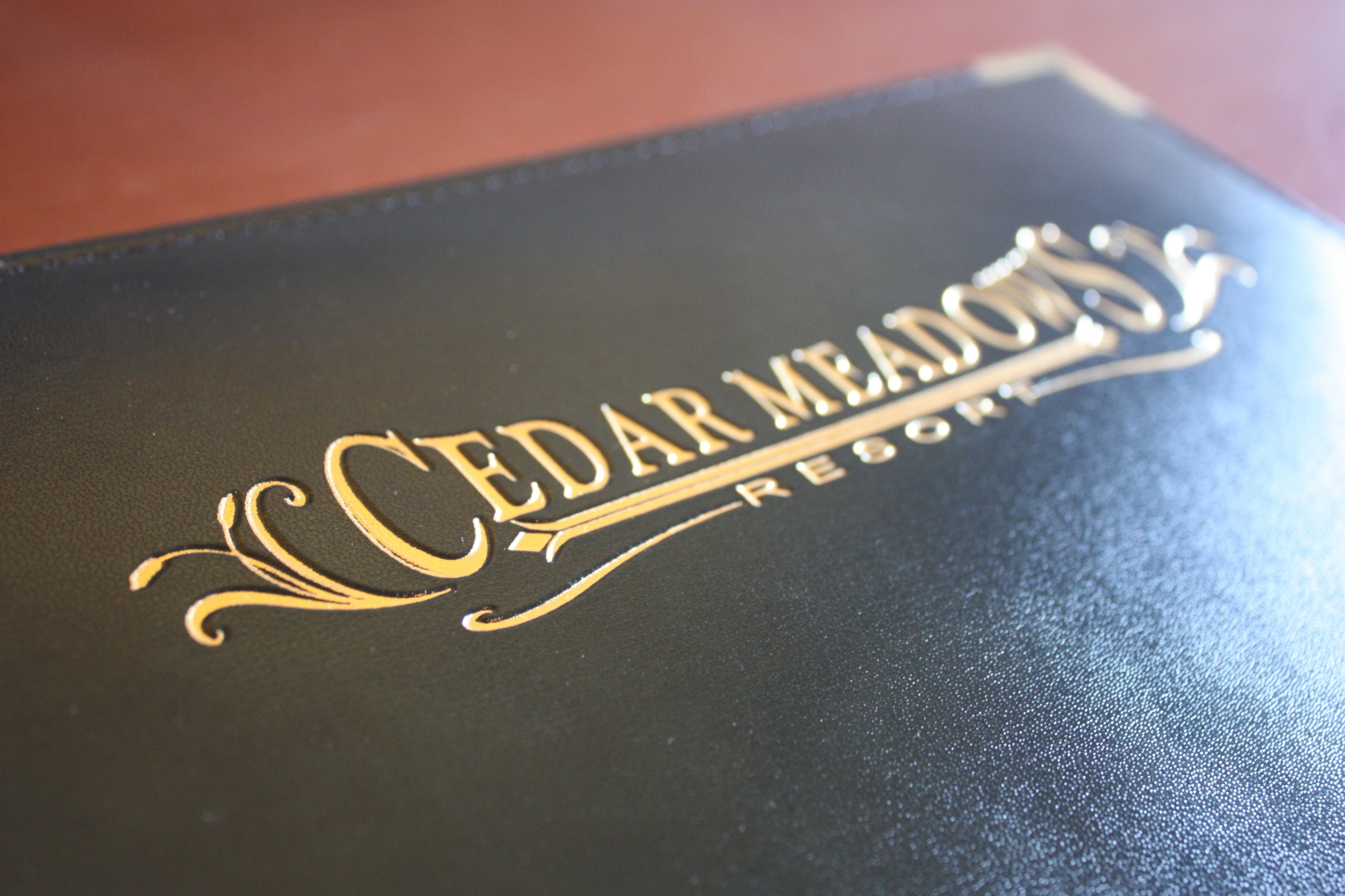 Un livre à la couverture en cuir noir, sur laquelle est estampillé en lettres dorées le texte « CEDAR MEADOWS RESORT ».