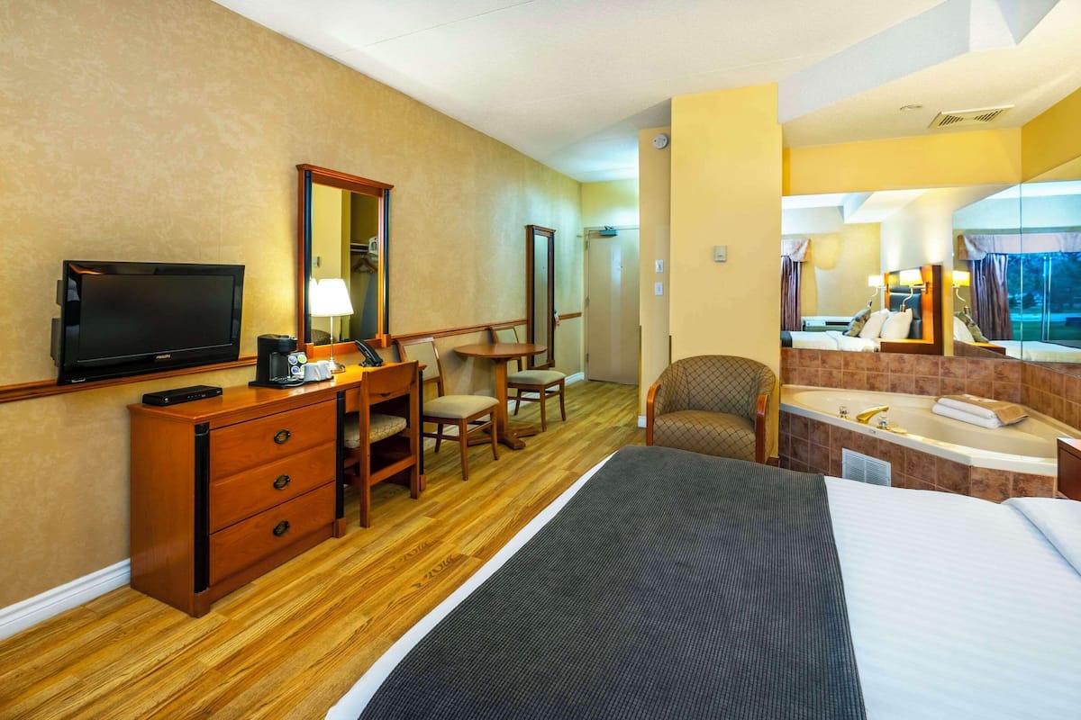 A hotel room with a bed, a desk with a TV, a chair, a small table, a mirror, and a bathroom with a bathtub and sink.