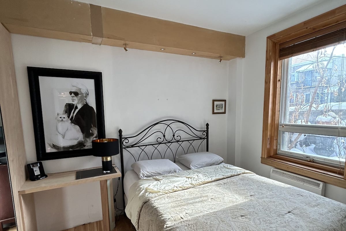 Une chambre à coucher avec un lit, une table de chevet avec une lampe, un tableau encadré au mur et une fenêtre donnant sur l’extérieur.