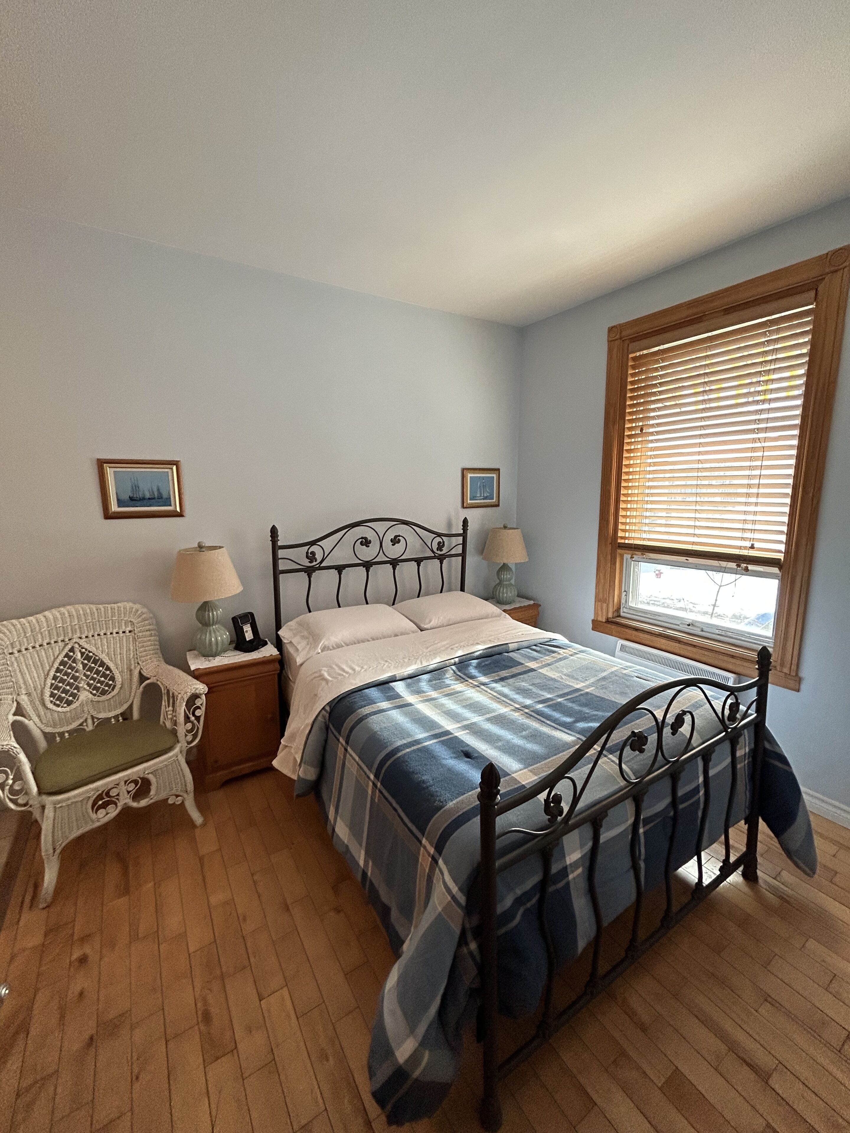 Une chambre à coucher avec un lit en métal, une chaise blanche avec un coussin, deux tables de chevet avec des lampes et une fenêtre avec des stores.