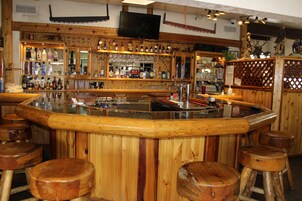 Zona de bar con taburetes de madera, una barra central y estantes llenos de botellas y copas.