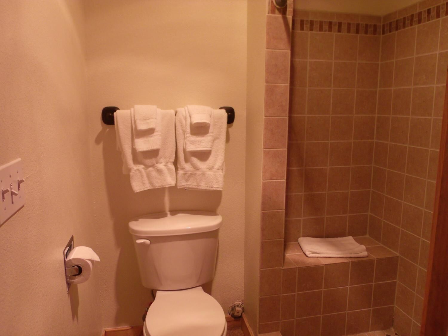 A bathroom with a toilet, two towels hanging on a holder, a shower area with tiled walls, and a toilet paper holder.