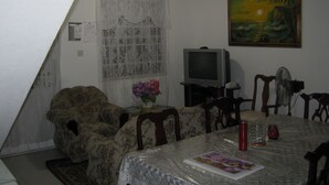 Una sala de estar con televisor, sillón floral, mesa de comedor con sillas, un jarrón con flores y un cuadro en la pared.