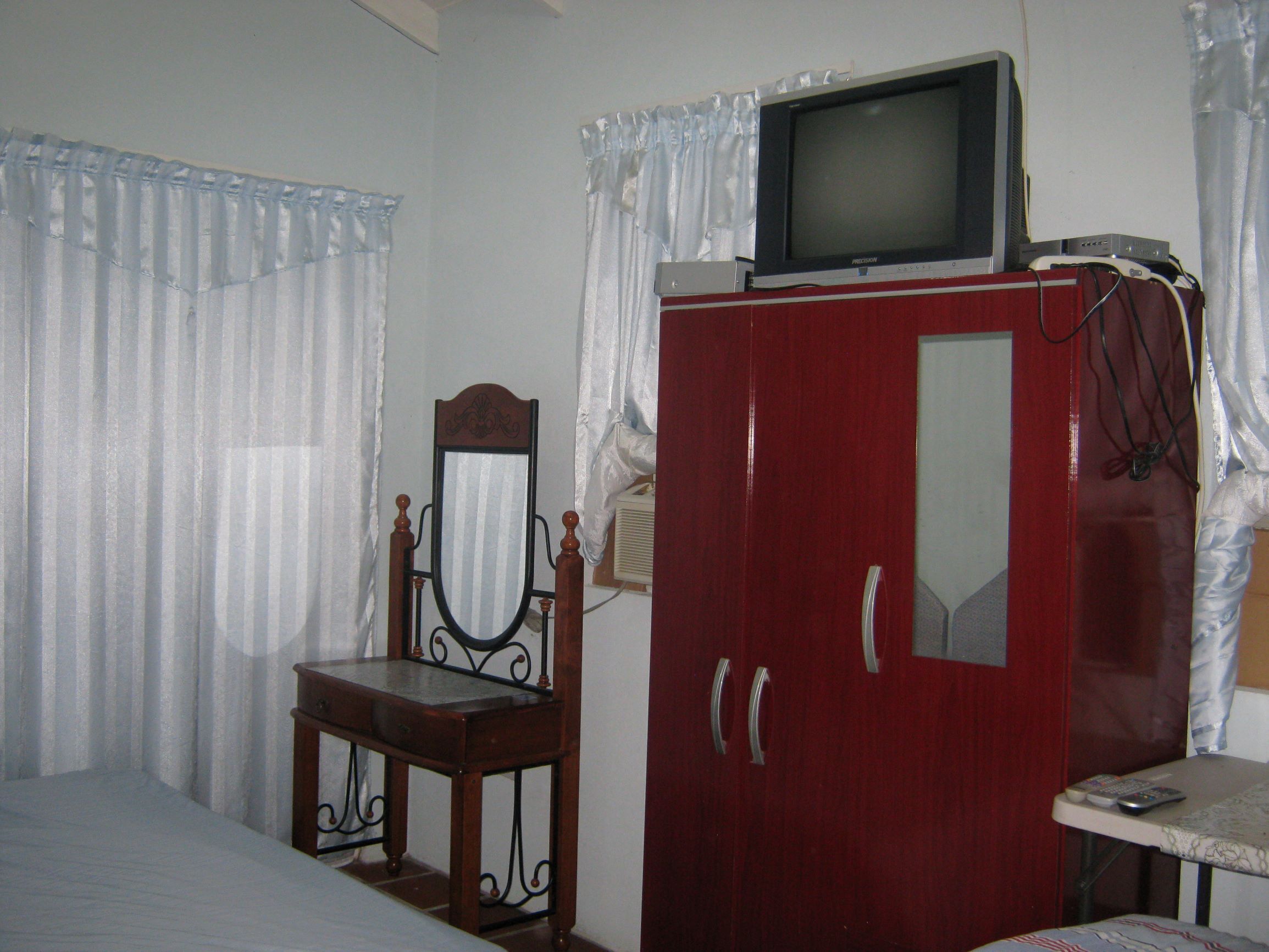 A bedroom with a bed, a wooden chair, a red wardrobe with a television on top, and a small table with a remote control.