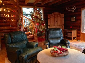 Interior acogedor de una cabaña de madera con dos sillones de cuero, una mesa de comedor de madera con un centro de frutas, un árbol de Navidad y una cabeza de venado montada en la pared.