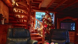 A cozy log cabin interior with two leather armchairs, a wooden staircase, a decorated Christmas tree, and a television in the background. - Handsome Dan's at Otter Point (Juan de Fuca Electoral Area)