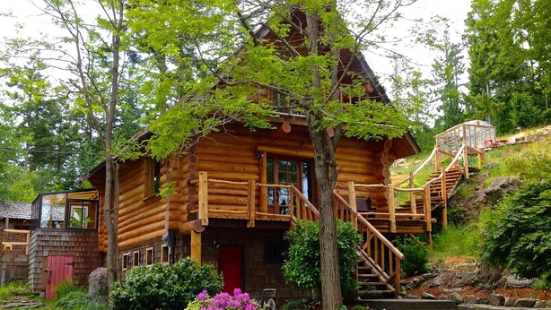 A log cabin with a wooden staircase leading to a porch, surrounded by lush greenery and a small garden with purple flowers. - Handsome Dan's at Otter Point (Juan de Fuca Electoral Area)