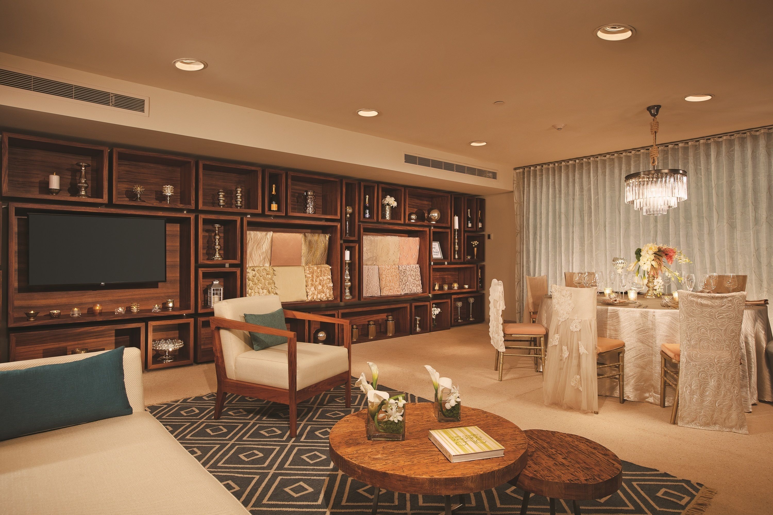 A spacious living room with a large flat-screen TV, comfortable seating, a dining area with a table set for two, and a decorative chandelier.