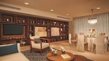 A spacious living room with a large flat-screen TV, comfortable seating, a dining area with a table set for two, and a decorative chandelier.