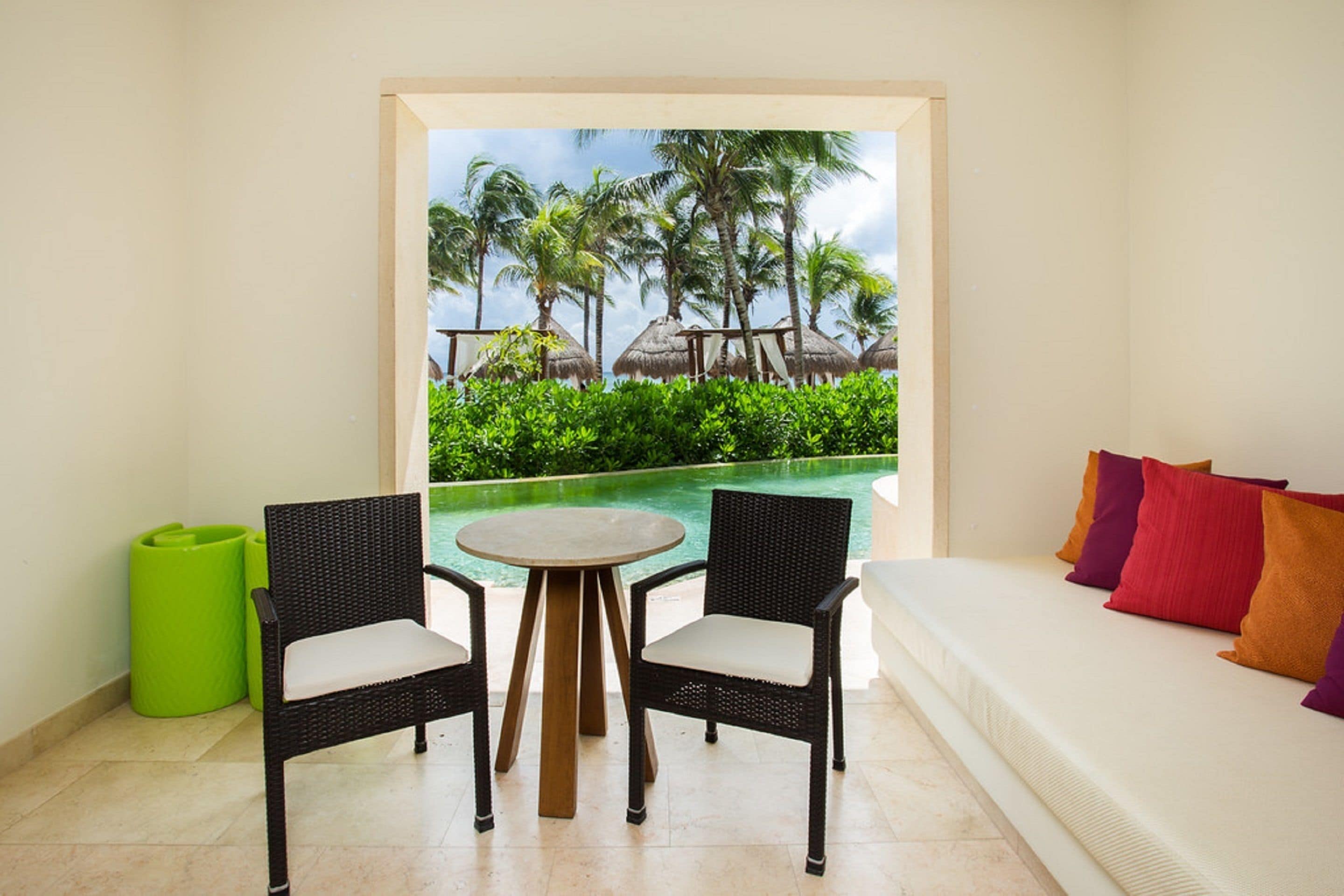 A room with a white sofa, two wicker chairs, a small round table, and a view of a pool with palm trees.