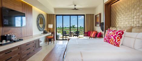 A modern hotel room with a large bed, a TV, a desk with a chair, and a balcony with a view of the outdoors.