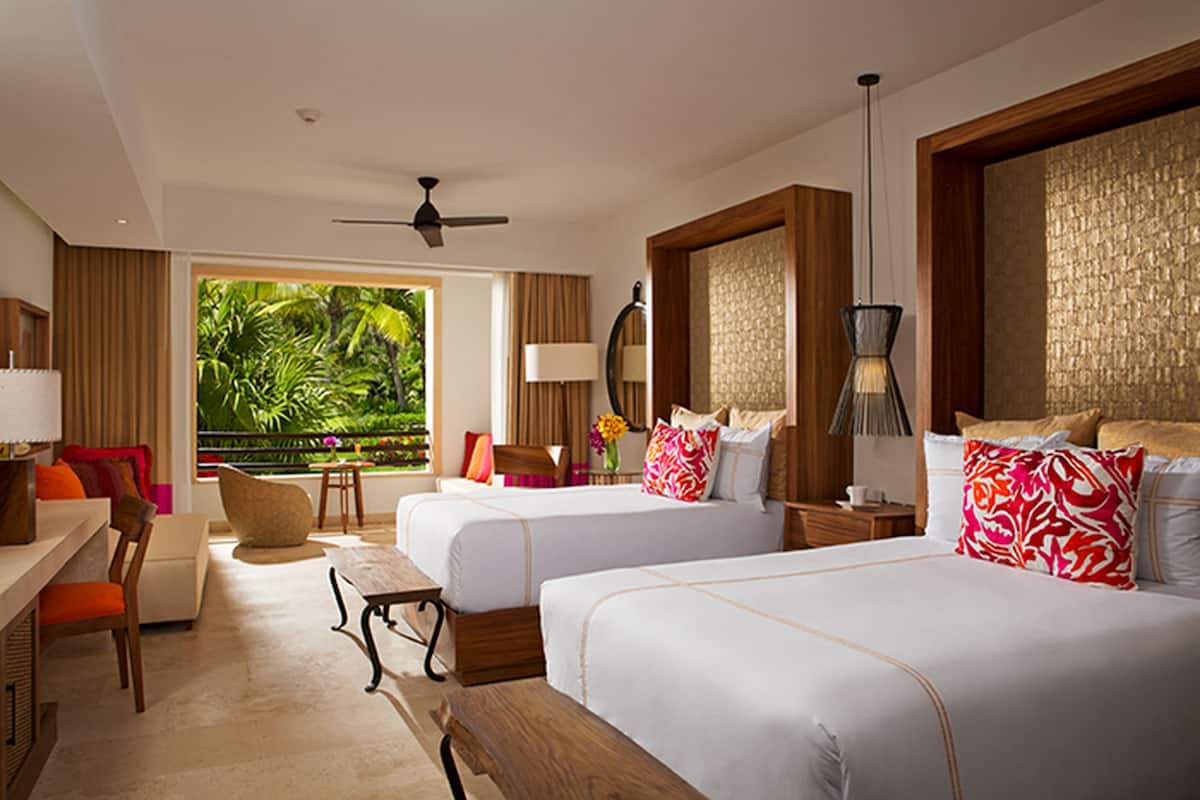 Quarto de hotel com duas camas, uma escrivaninha, uma cadeira, varanda com vista para área verde e um ventilador de teto.