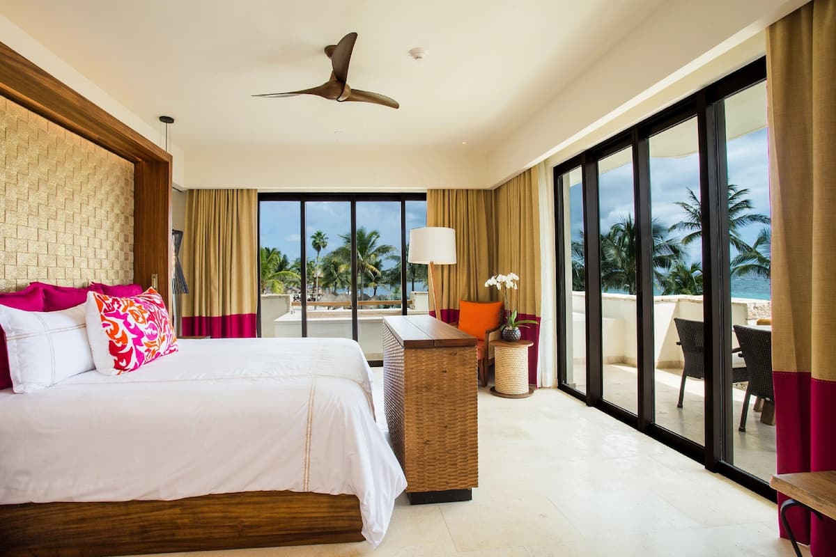 Quarto de hotel com uma cama grande, ventoinha no teto, vista para palmeiras e varanda com área de estar.