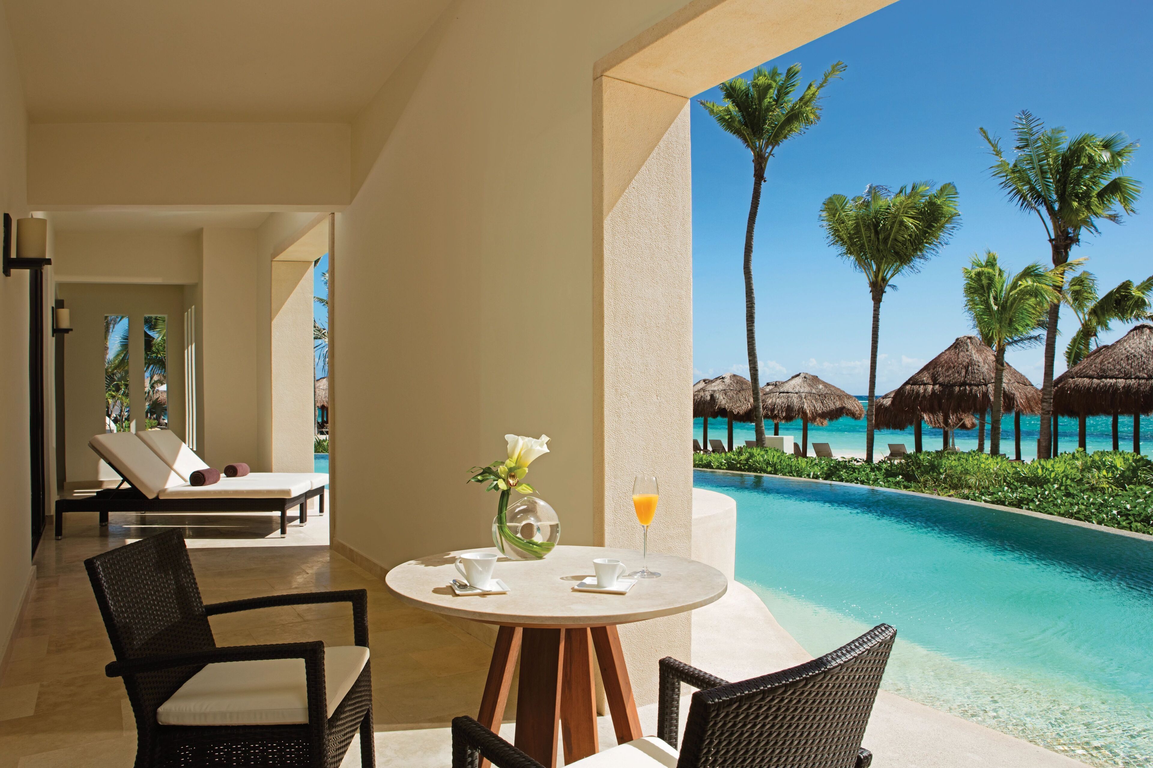 A poolside area with lounge chairs, a table with a vase and a glass of orange juice, and a view of the ocean with palm trees and thatched umbrellas.