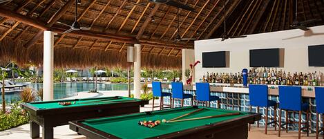 A pool area with two pool tables, a bar with chairs, and a thatched roof.