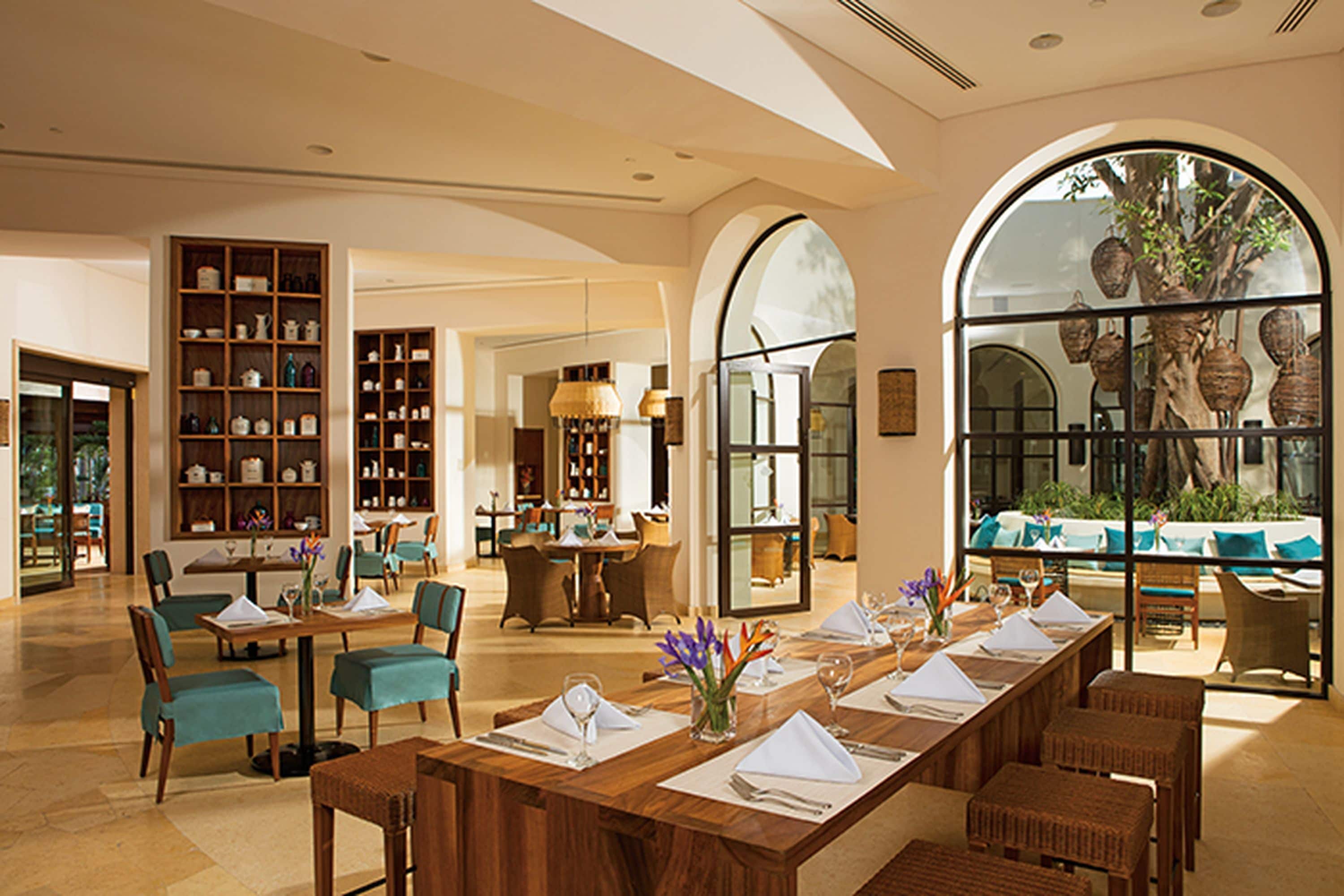 A spacious hotel lobby with a long dining table set for a meal, surrounded by comfortable seating and a large arched window offering a view of the outdoors.