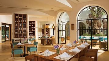 A spacious hotel lobby with a long dining table set for a meal, surrounded by comfortable seating and a large arched window offering a view of the outdoors.
