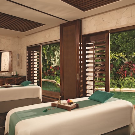 A spa room with two beds, a stone wall, a large window, and a view of greenery.