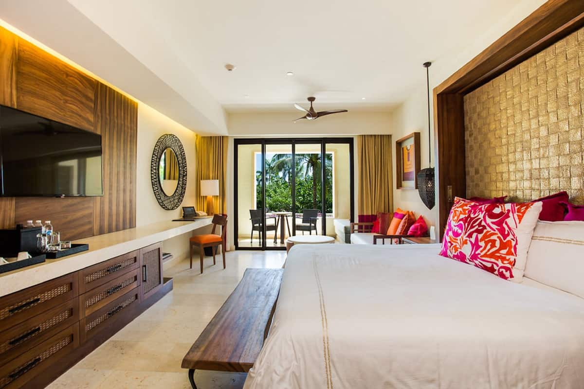 Quarto de hotel com uma cama grande, uma escrivaninha com televisão, varanda com cadeiras e ventoinha no teto.