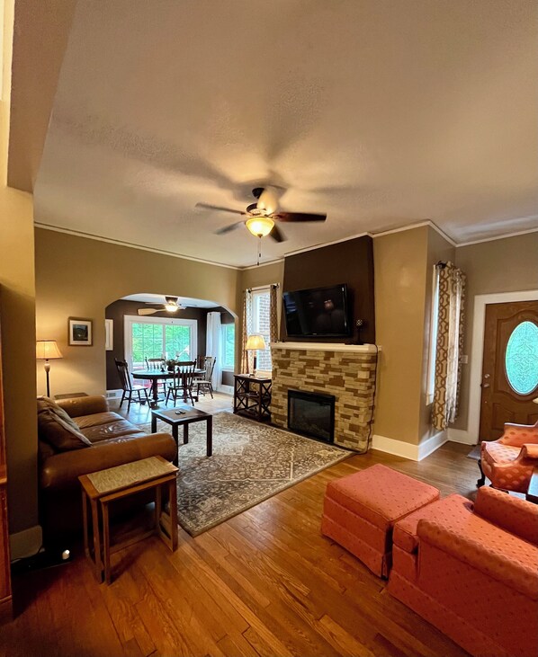 A living room with a fireplace, a ceiling fan, a television, and a dining area with a table and chairs. - Grafton Lodge (Lake Lure)