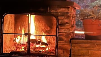 A stone fireplace with a fire burning inside, surrounded by logs and a brick floor.