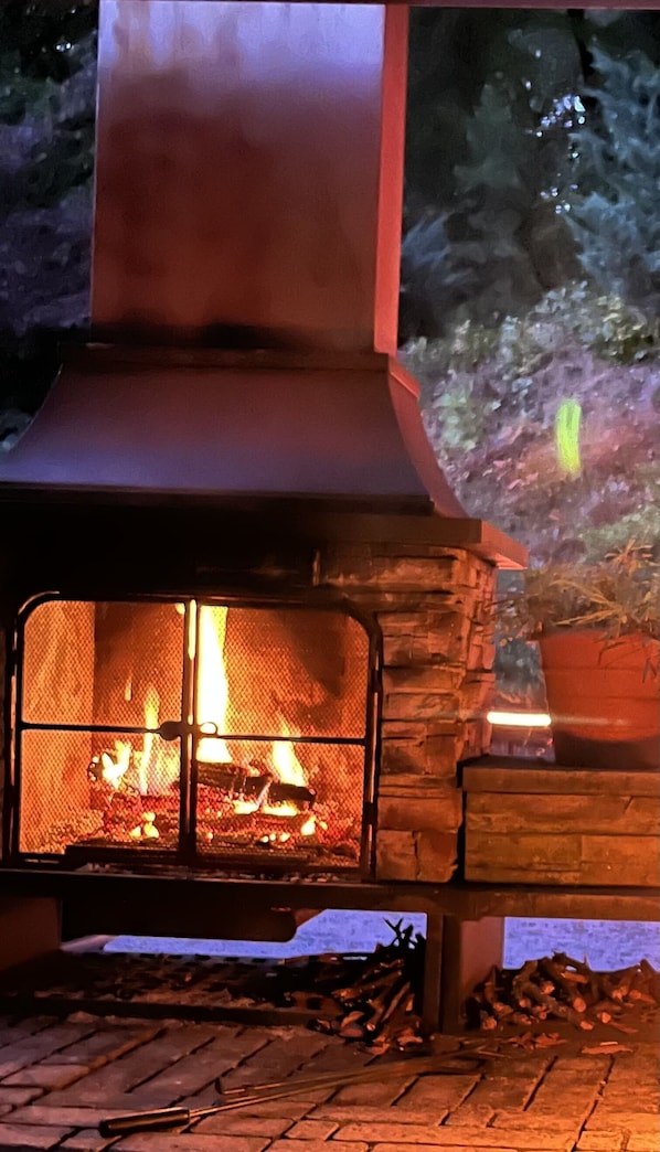 A stone fireplace with a fire burning inside, surrounded by logs and a brick floor.