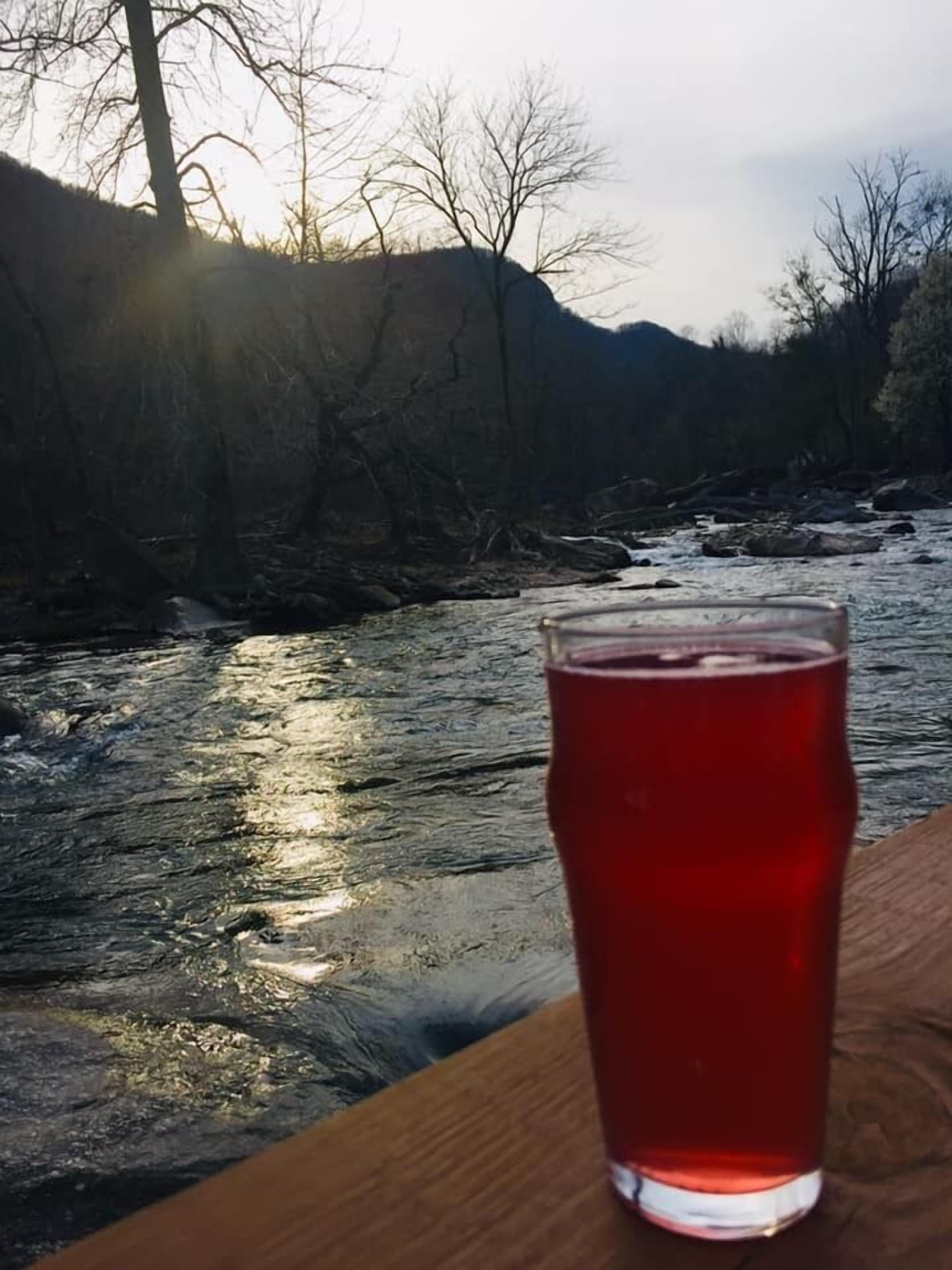 Un vaso de bebida roja sobre una superficie de madera junto a un río, con árboles desnudos y montañas al fondo.