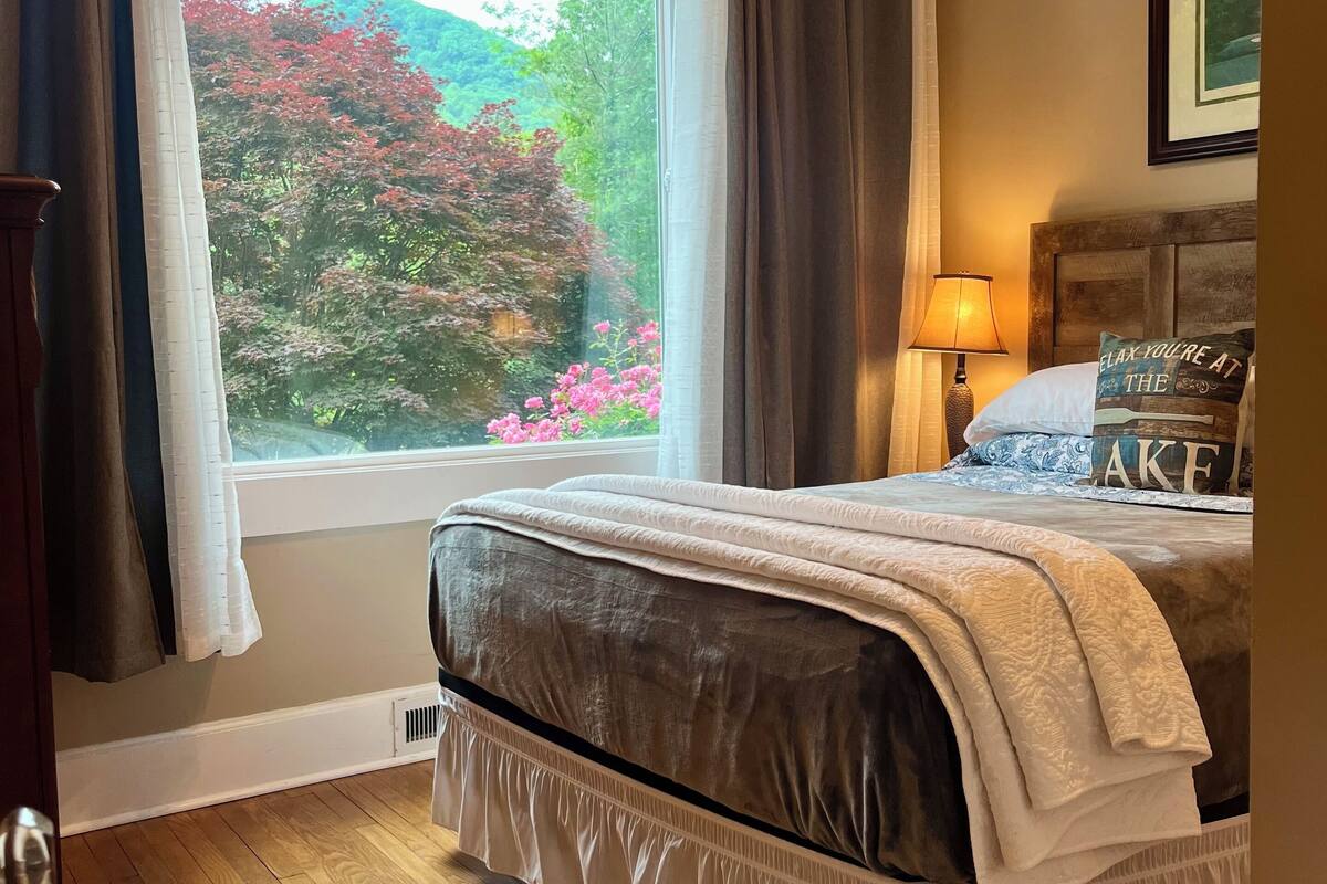 A bedroom with a large window, a bed with a headboard, a bedside lamp, and a framed picture on the wall.
