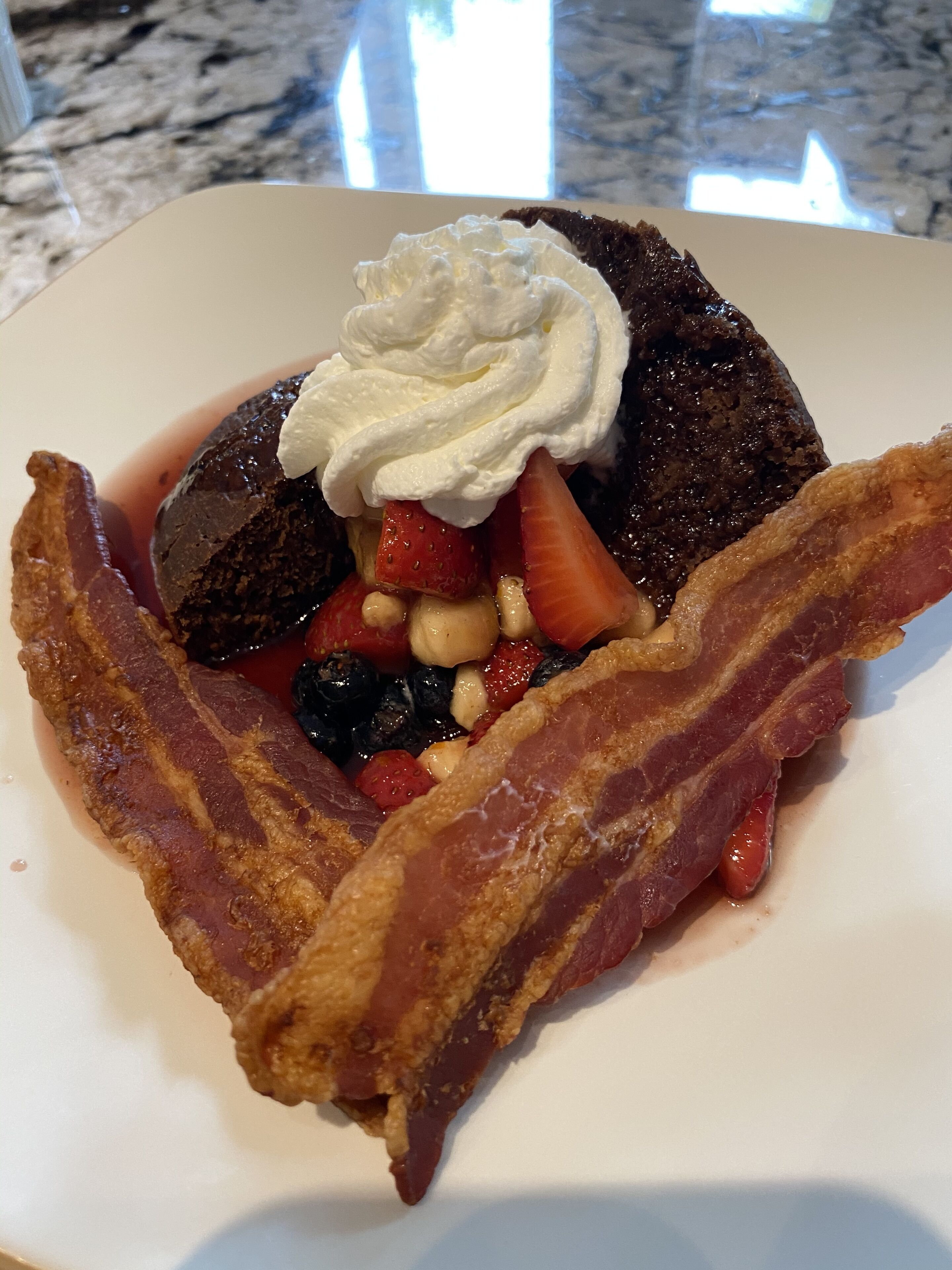 Um prato com sobremesa de chocolate coberta com chantilly e frutas vermelhas frescas, acompanhado de fatias crocantes de bacon.