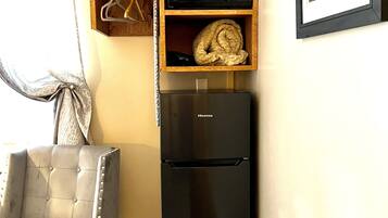 Un espace de vie compact comprenant un réfrigérateur Hisense noir, une étagère en bois avec une plante et une serviette enroulée, un fauteuil gris à motifs et un tableau encadré accroché au mur.