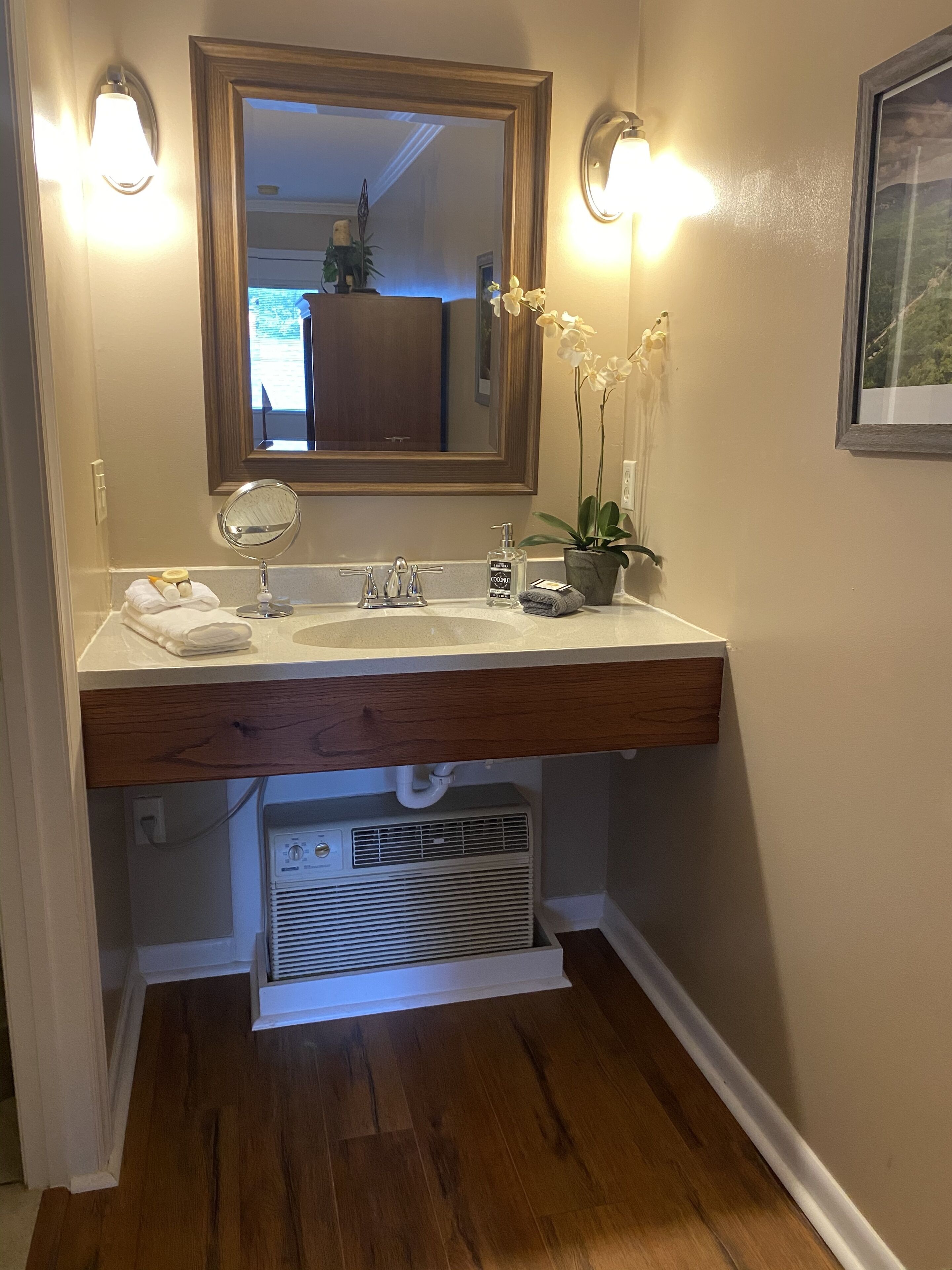 Une salle de bain avec un meuble-lavabo en bois, un grand miroir, deux appliques murales, une orchidée en pot et un tableau encadré accroché au mur.
