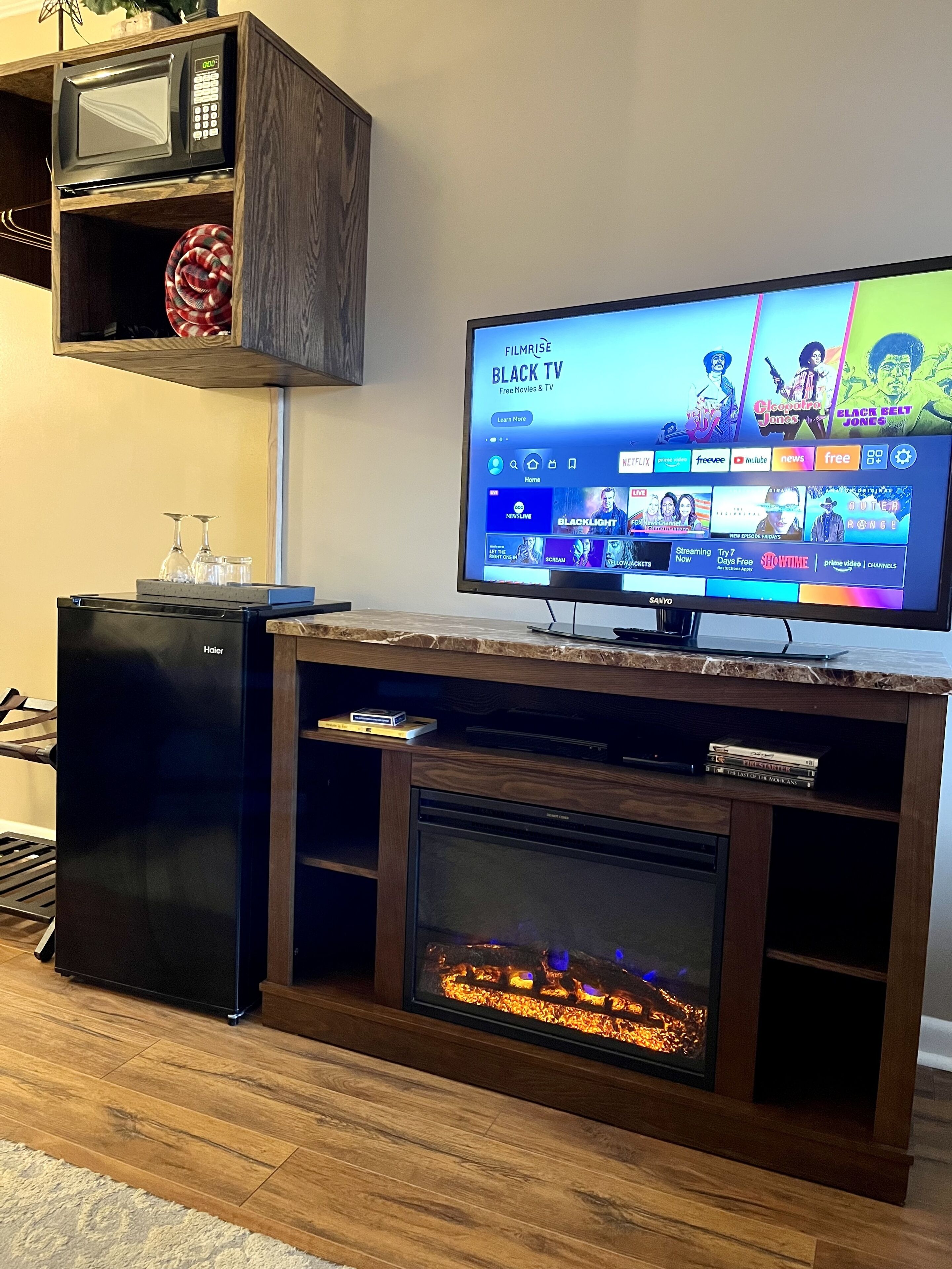 Una sala de estar con televisión de pantalla plana sobre un mueble de madera, un refrigerador pequeño y una chimenea empotrada.