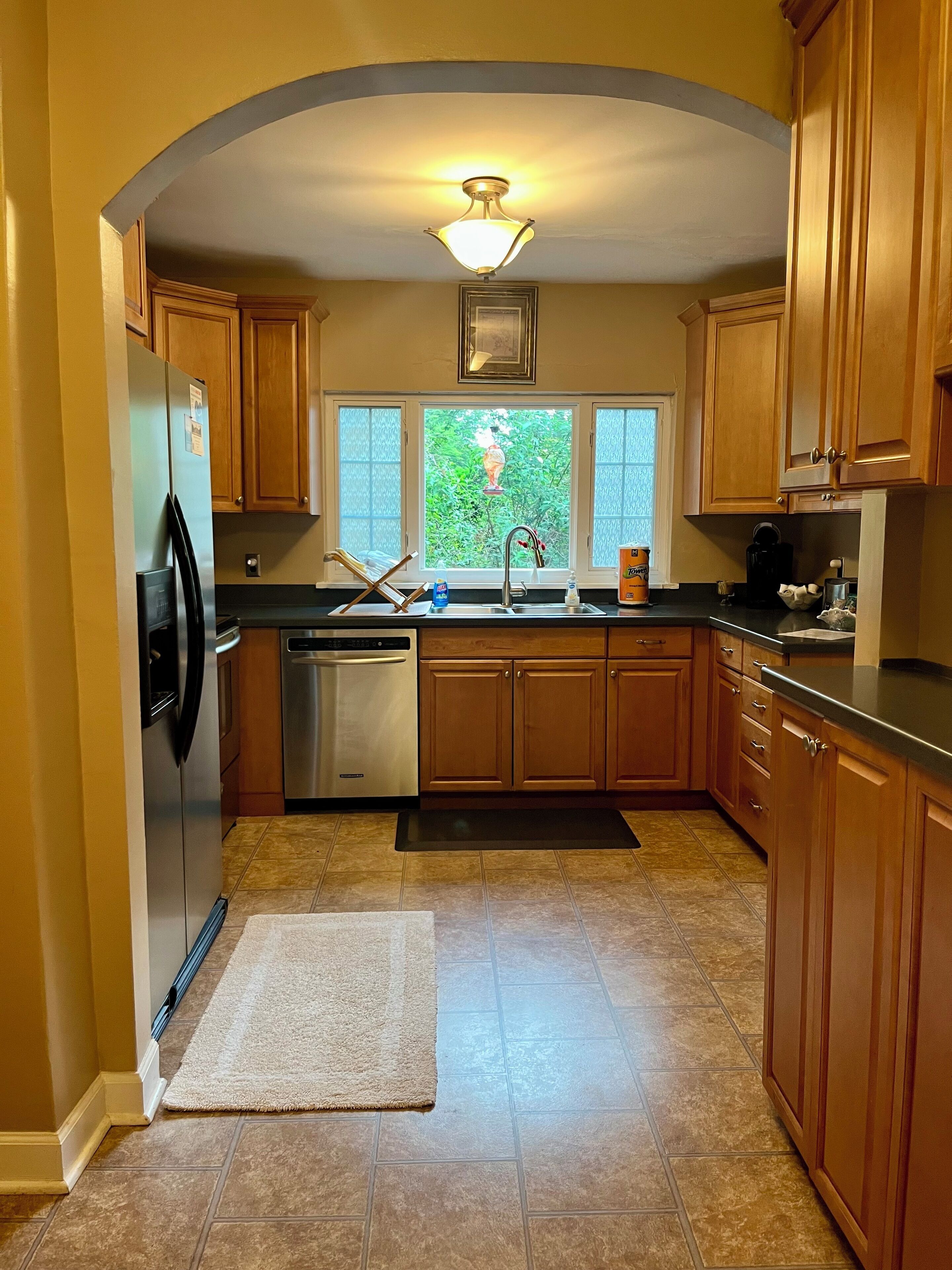A kitchen with wooden cabinets, a stainless steel refrigerator, a dishwasher, and a window with a view of greenery.