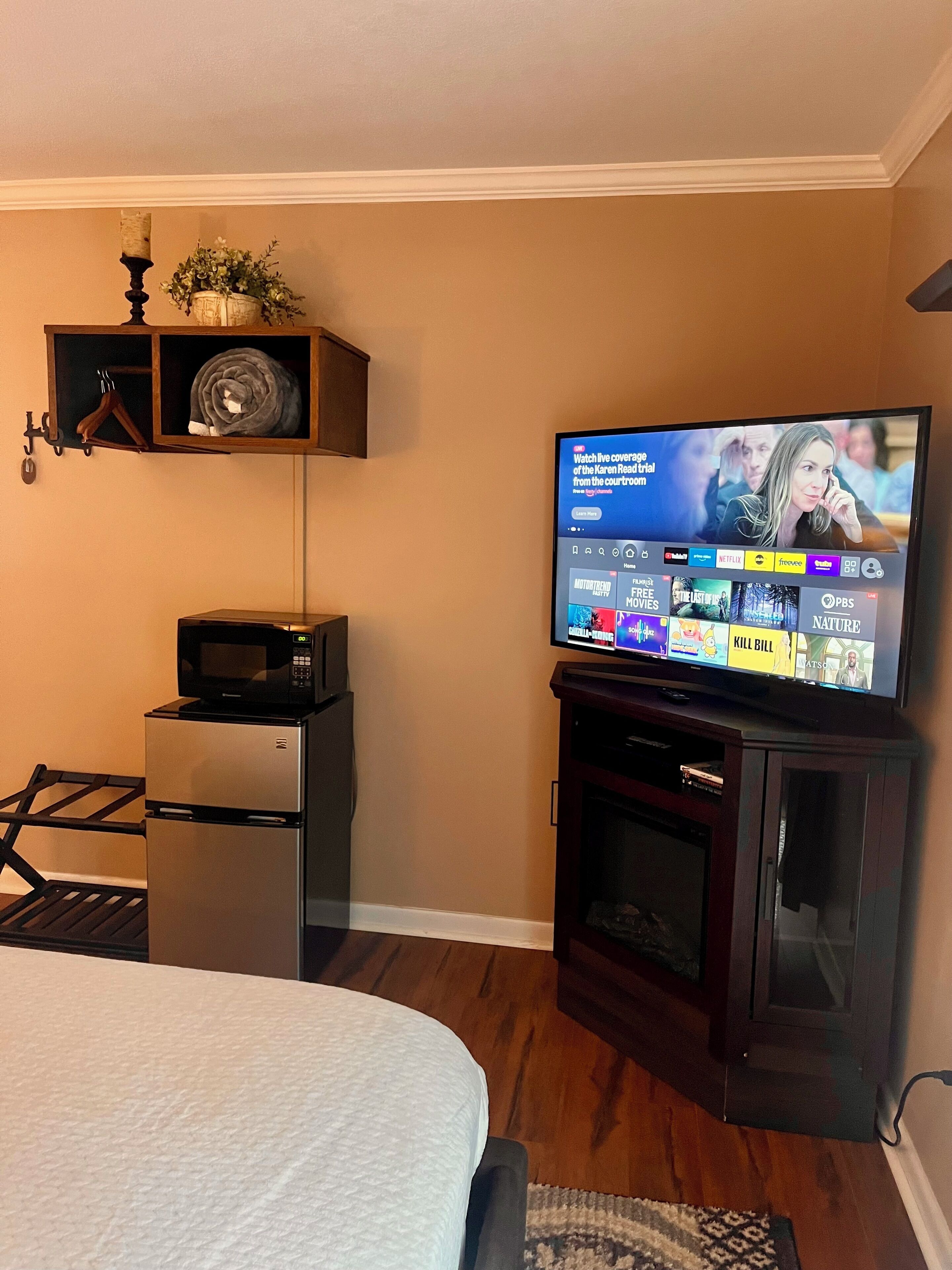 Une chambre d’hôtel équipée d’un téléviseur à écran plat diffusant une chaîne d’information, d’un four à micro-ondes, d’un réfrigérateur, d’un lit et d’une petite armoire contenant une plante et une bougie.