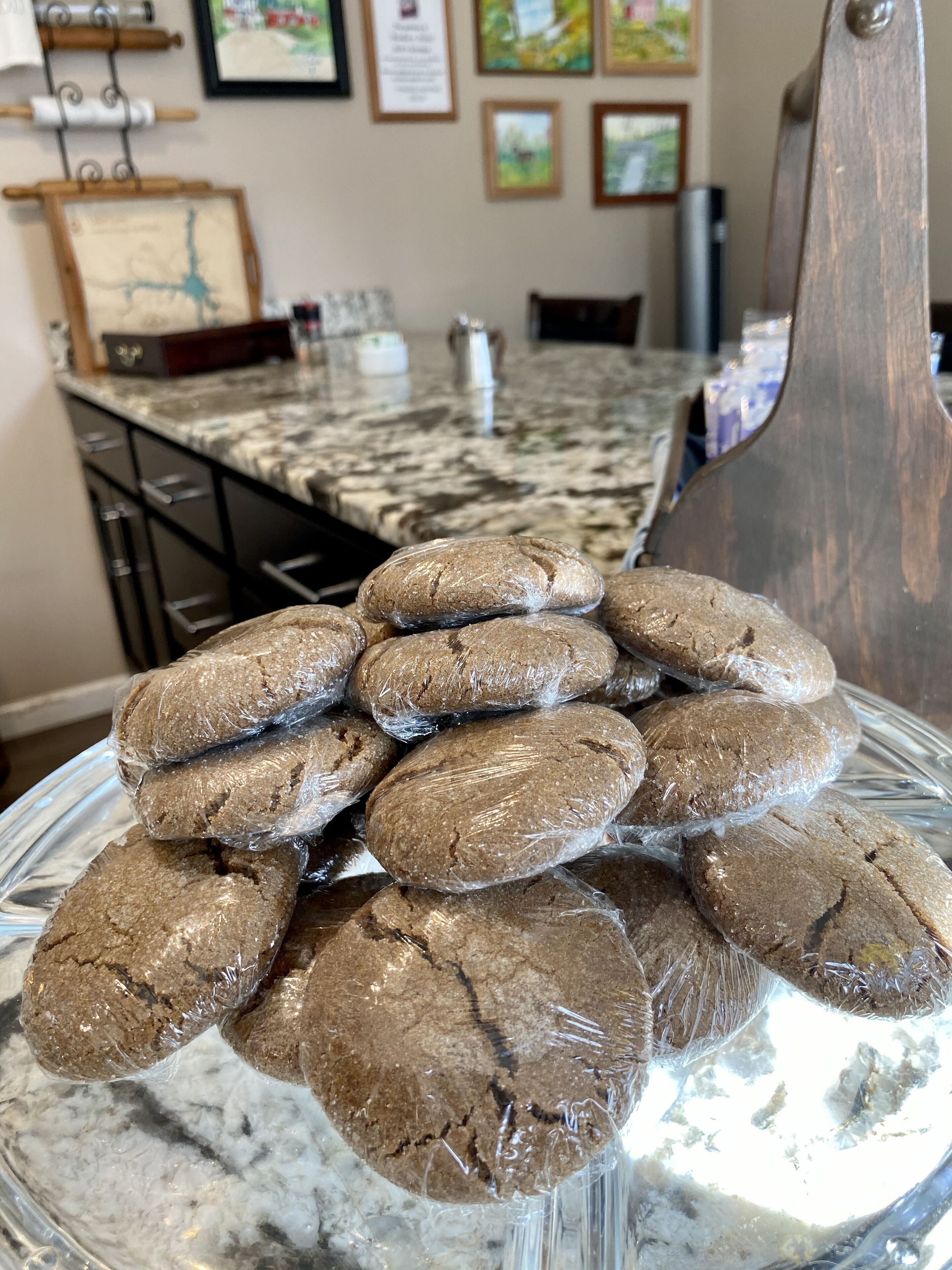 Um prato de vidro com vários biscoitos redondos marrons, embalados em plástico, sobre um balcão de mármore em uma cozinha.