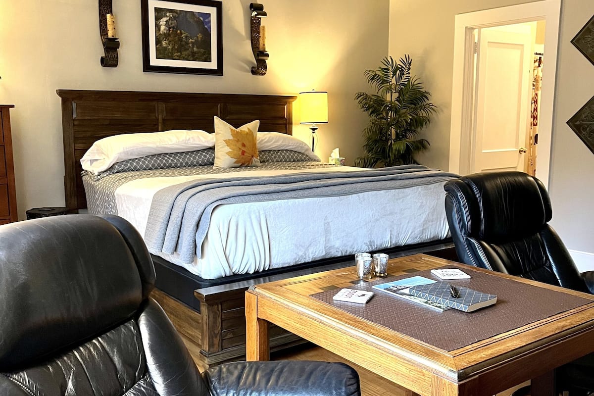 A hotel room with a bed, two black leather chairs, a wooden table, a nightstand, a lamp, a framed picture, and a potted plant.