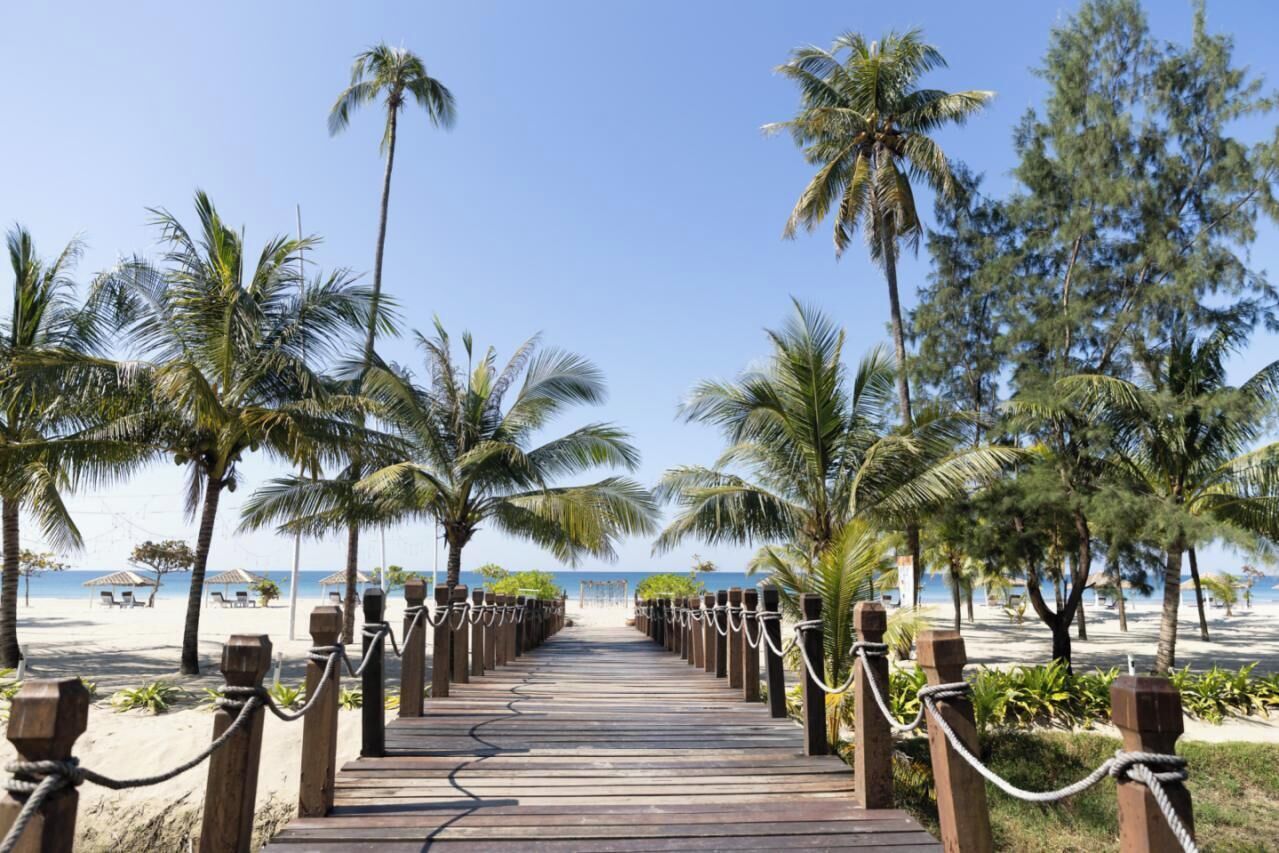 A wooden boardwalk leading to a beach with palm trees and beach chairs.
